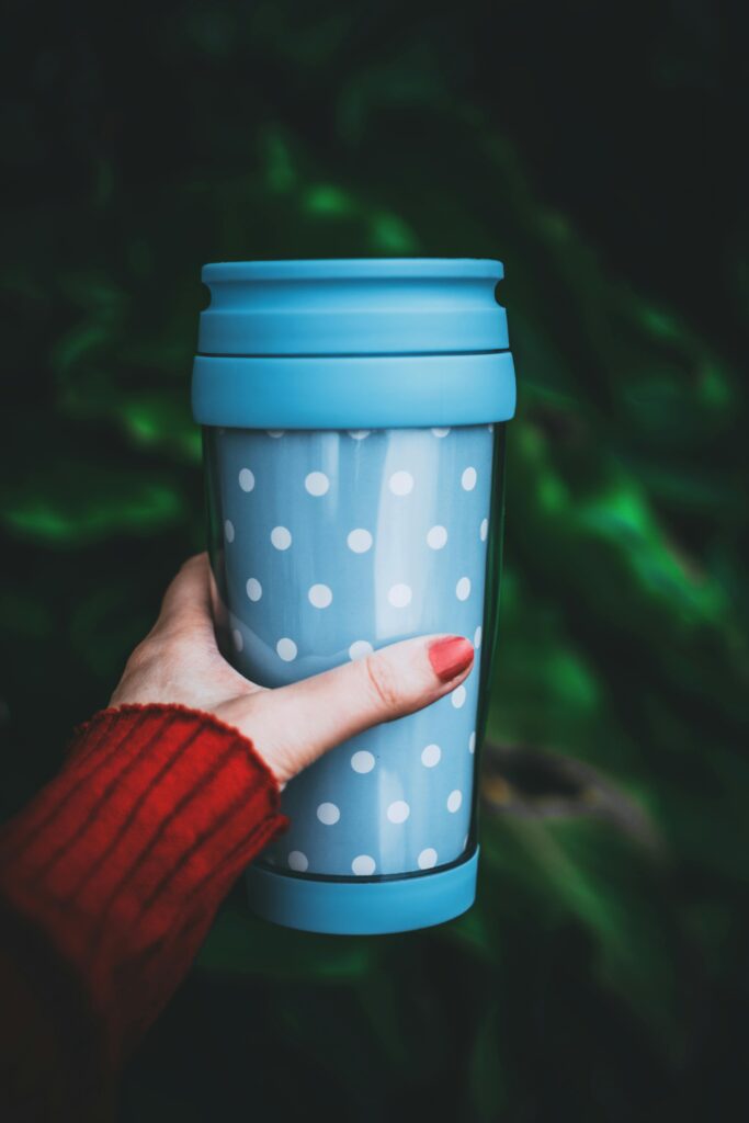 A close-up of a person holding a blue polka dot travel mug against a green background.