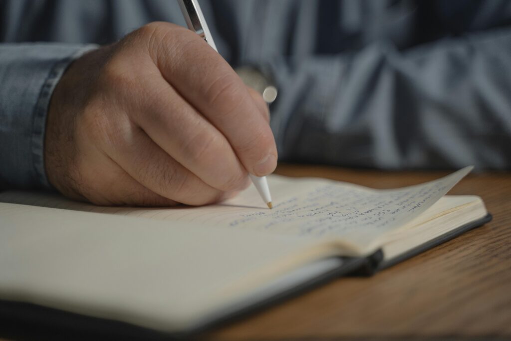 A close-up image of a hand writing with a pen in a notebook, creating a personal diary entry.